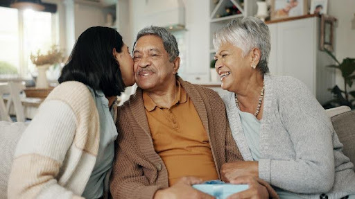 A daughter kissing her father on the cheek, sitting on a couch with an older woman beside them.