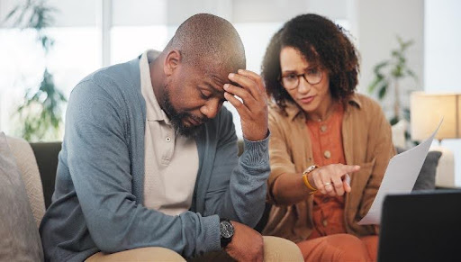 A man sits with his head resting on his hand, while a woman beside him points to paperwork.