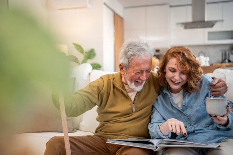 Senior man with walking cane and his young adult granddaughter are sitting on a sofa at home, looking at a photo album together and laughing, while the granddaughter is holding a mug